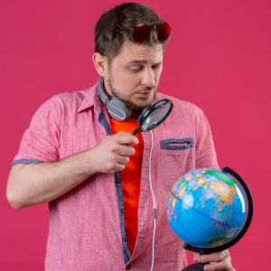 young traveler man with headphones looking globe through magnifying glass standing pink background