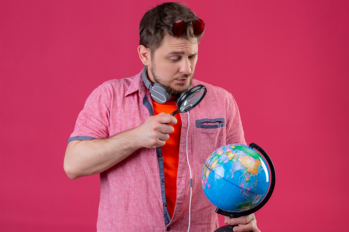 young traveler man with headphones looking globe through magnifying glass standing pink background
